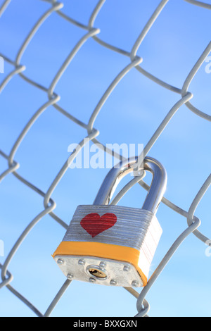 Locked padlock with heart hanging on a chain linked fence with a blue sky in the background. Stock Photo