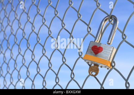 Unlocked padlock with heart and key hanging on a chain linked fence with a blue sky in the background. Stock Photo