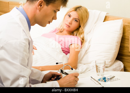 Sick female lying in bed and looking at male doctor while he prescribing her medication Stock Photo