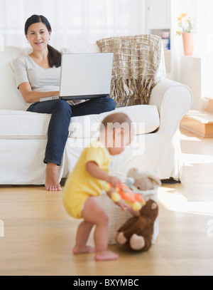 Baby boy playing, mother sitting on sofa and using laptop Stock Photo
