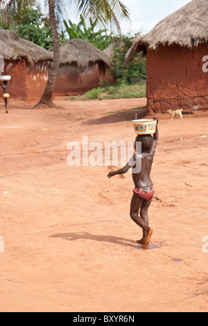 A young African child living in rural Togo balances water on her head while transporting it from the village well to her home. Stock Photo