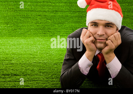 Happy guy in suit and santa hat lying on green grass and touching his face smiling Stock Photo