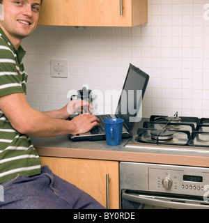 Young single man, 20s, in kitchen drinking coffee and working at laptop computer, smiling at camera Stock Photo