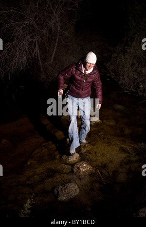 A women dressed in white hat, purple dawn jacket and light blue jeans crossing a river over stepping stones in the night. Stock Photo