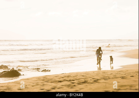 A boy rides his bicycle while a dog runs alongside on a sandy beach in Mexico. Stock Photo