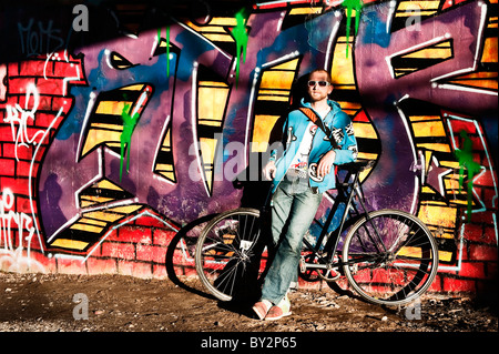 A man leaning against a graffiti covered wall in front of his bicycle. Stock Photo