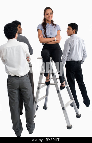 One businesswoman sitting on top of  a ladder  facing the camera and group of businessmen standing around her and staring Stock Photo