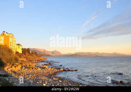 Bright morning sunlight shining on hotels, gravel beach, Lake Nahuel Huapi and Cerro Lopez Andean peaks, Bariloche, Argentina Stock Photo