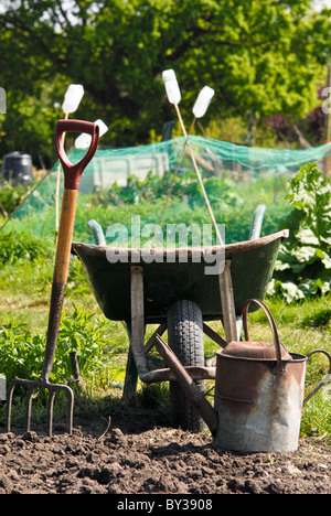 An old rusty fork, wheelbarrow and watering can on an allotment in early summer, with garden canes and trees in the background Stock Photo