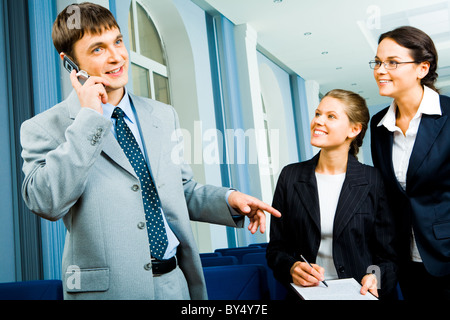 Portrait of busy man talking on the cell while two young pretty women standing near by and looking at him Stock Photo