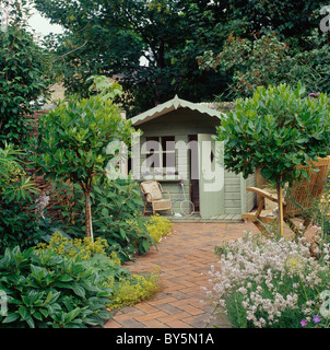 Standard bay trees on either side of brick paved path leading to pale green summer house in country garden in summer Stock Photo