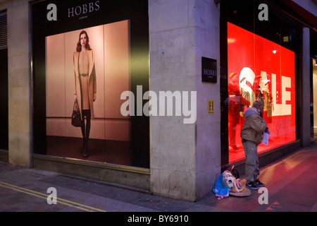 A Big Issue seller awaits custom with her pet dog on pavement outside clothing shop Hobbs in central London. Stock Photo