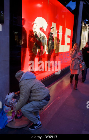 A Big Issue seller makes her pet dog comfortable on pavement outside clothing shop Hobbs in central London. Stock Photo