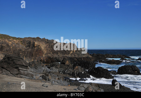 Blue sky beach view of white foam waves washing shiny black blocks of basalt in front of a cliff platform, Chiloe Island, Chile Stock Photo