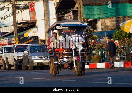 A colorful tuk-tuk with orange wheels is transporting a male passenger on a bright sunlit city street in Nong Kai, Thailand. Stock Photo