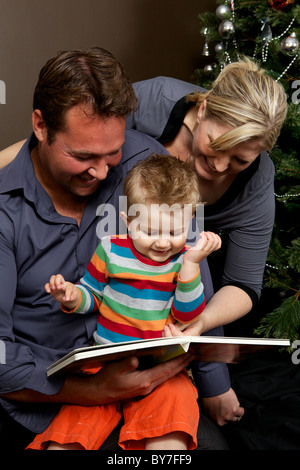 Mother and father reading a book together with their toddler son at Christmas time by the Christmas tree