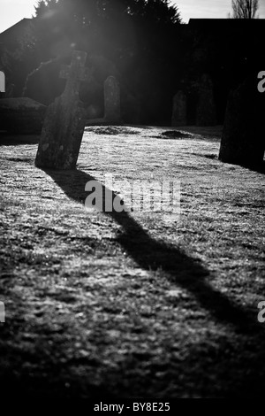 Black and white photo of a stone cross in a church graveyard.  The back light from the sun has created a cross shaped shadow Stock Photo