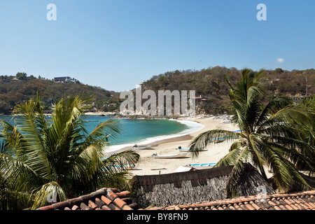 view looking down across tile & thatched roofs to crescent of beach & blue green water of Puerto Angel bay Oaxaca State Mexico Stock Photo