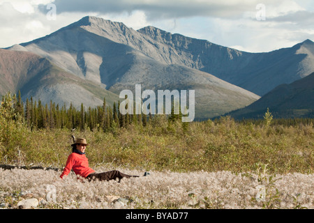 Young woman relaxing, enjoying evening light, sitting in the grass, Cotton Grass, Northern Mackenzie Mountains behind Stock Photo