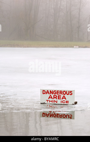 A pond covered in a thin layer of ice with some open water, and a sign signifying the danger during a foggy day. Stock Photo