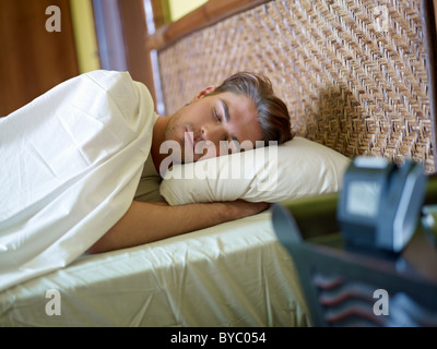 serene caucasian adult man lying in bed with alarm clock in foreground. Horizontal shape, waist up, front view Stock Photo