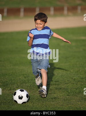 Authentic happy Latino boy playing with soccer ball in field wearing blue striped tee shirt. Stock Photo