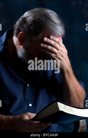 Man holds his head in one hand and bows to pray while holding his bible in the other. Stock Photo