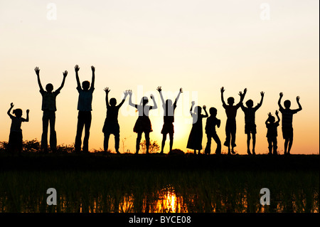 Silhouette Indian girls and boys jumping and waving standing on a rice paddy field at sunset. Andhra Pradesh, India Stock Photo