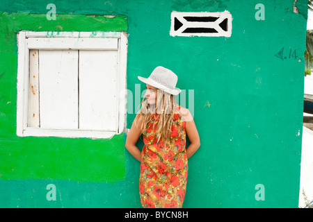Woman with blond dreadlocks in red dress and white hat leaning against a green wall in Panama Stock Photo