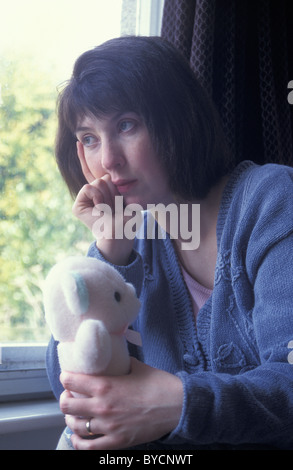 unhappy woman sitting by window holding child's teddy Stock Photo