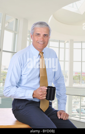 Middle aged businessman sitting on the corner of his desk holding a coffee mug. Stock Photo