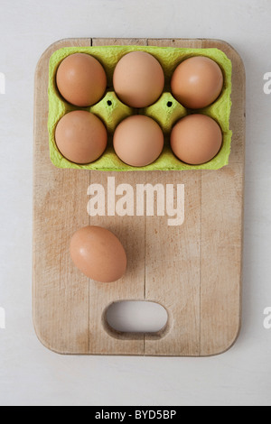 Six brown eggs in an egg carton and one egg on a wooden cutting board beside it Stock Photo