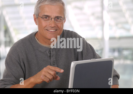 Middle aged man in lobby of modern office building laptop computer. Man is casually dressed and smiling and pointing at computer Stock Photo