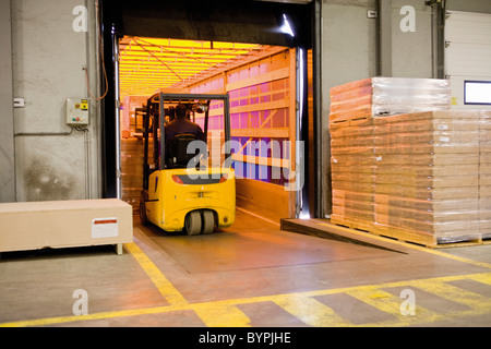 Forklift operator loading wrapped pallets of cardboard boxes onto trailer in warehouse Stock Photo