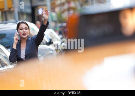 A young woman or businesswoman hailing a yellow taxi cab while talking on her cell phone in a modern city Stock Photo