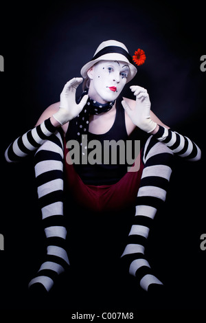 Studio portrait of Dramatic funny mime in white hat with red flower and striped gloves sitting on floor on black background Stock Photo