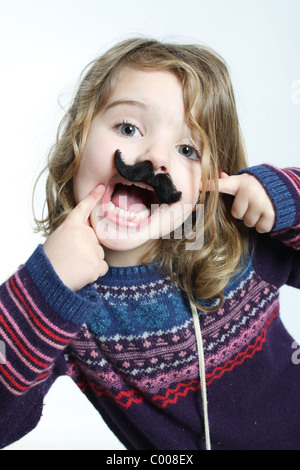 A little white caucasian girl pointing  at her fake black moustache, happy with mouth open. Stock Photo