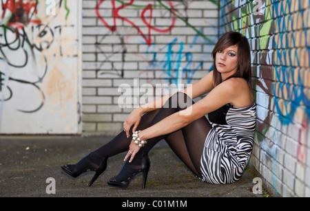 Young woman with dark hair wearing a top with a zebra pattern and high heels posing while sitting in front of a wall with Stock Photo