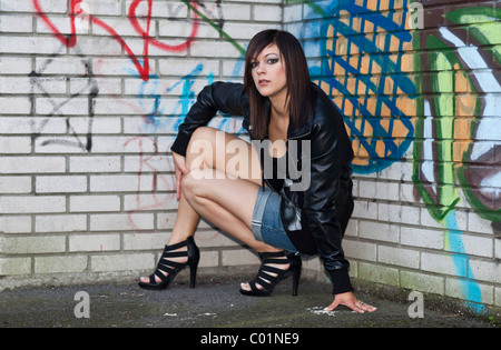 Dark-haired young woman wearing hot pants, a black leather jacket and high heels posing in front of a wall with graffiti Stock Photo
