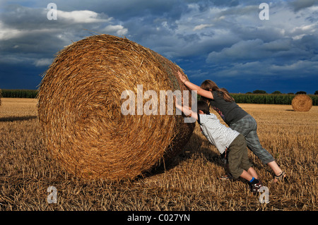 Little boy, six years, and girl, 10 years, trying to push a bale of hay on a stubble field, in the back a stormy sky Stock Photo