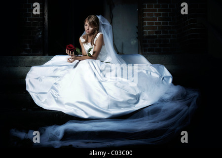 Young woman in a wedding dress and veil, sitting on a staircase in an urban location and holding a red rose in her hand Stock Photo