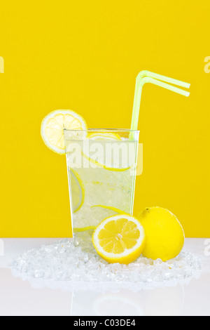 Photo of traditional lemonade in a glass with crushed ice and lemon slices, on a white table with yellow background. Stock Photo