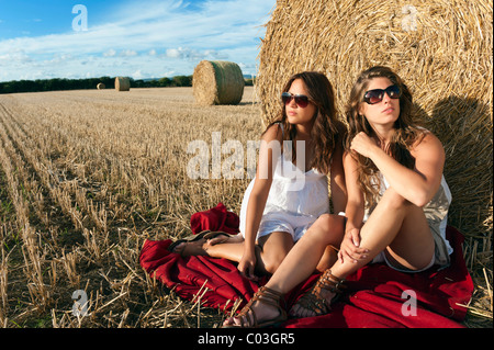 Two young women sitting on a harvested grain field in front of a bale of straw and talking, Finistere departement Stock Photo