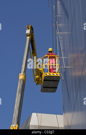 Worker wearing hard hat standing on an articulated telescoping boom lift near the top of a facade, worm's-eye view Stock Photo