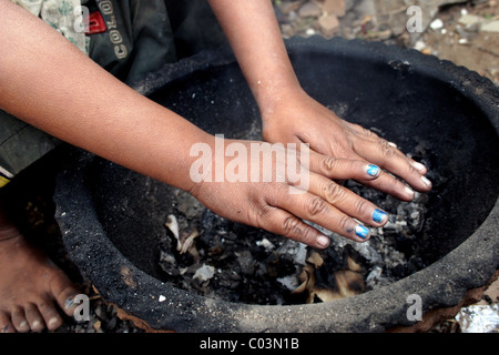 A young 9 year old child laborer boy living in poverty is warming his hands over a wood burning stove in Cambodia. Stock Photo