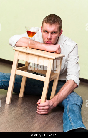 Drunken man sitting on floor with glass of alcohol and holding stool Stock Photo