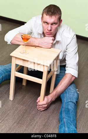 Drunken man sitting on floor with glass of alcohol and holding stool Stock Photo