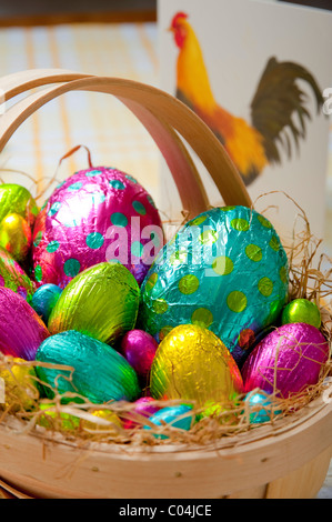 Traditional wooden basket filled with colorful foil wrapped chocolate easter eggs on yellow and white tablecloth Stock Photo