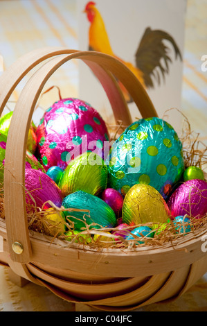 Traditional wooden basket filled with colorful foil wrapped chocolate easter eggs on yellow and white tablecloth Stock Photo