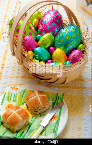 wooden basket filled with colorful foil wrapped chocolate easter eggs on yellow and white tablecloth Hot cross buns on plate Stock Photo